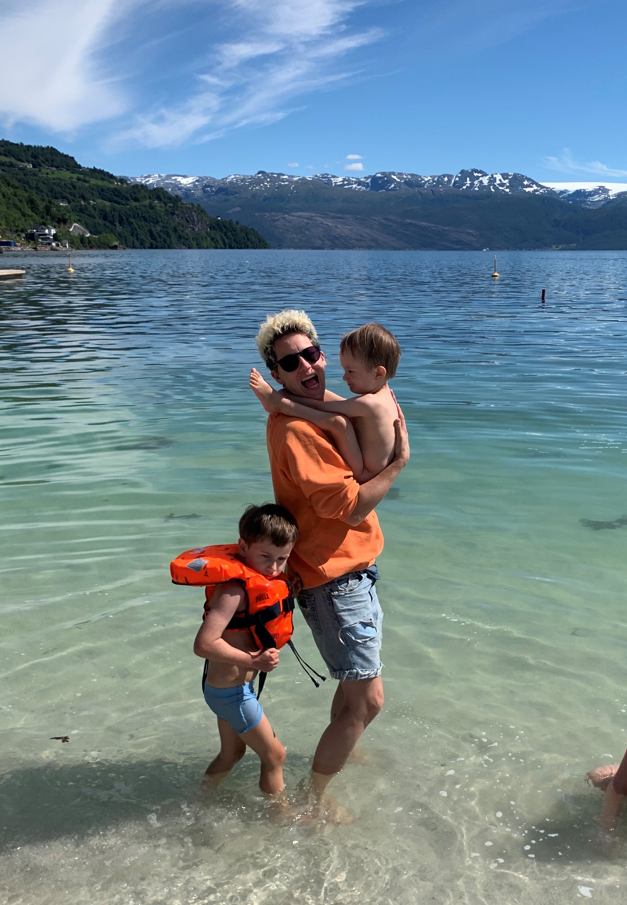 Heidi Marie vasser i havet, hun har solbriller på og oransje genser. Hun bærer på sønnen sin og ved siden av henne er den andre sønnen med redningsvest.