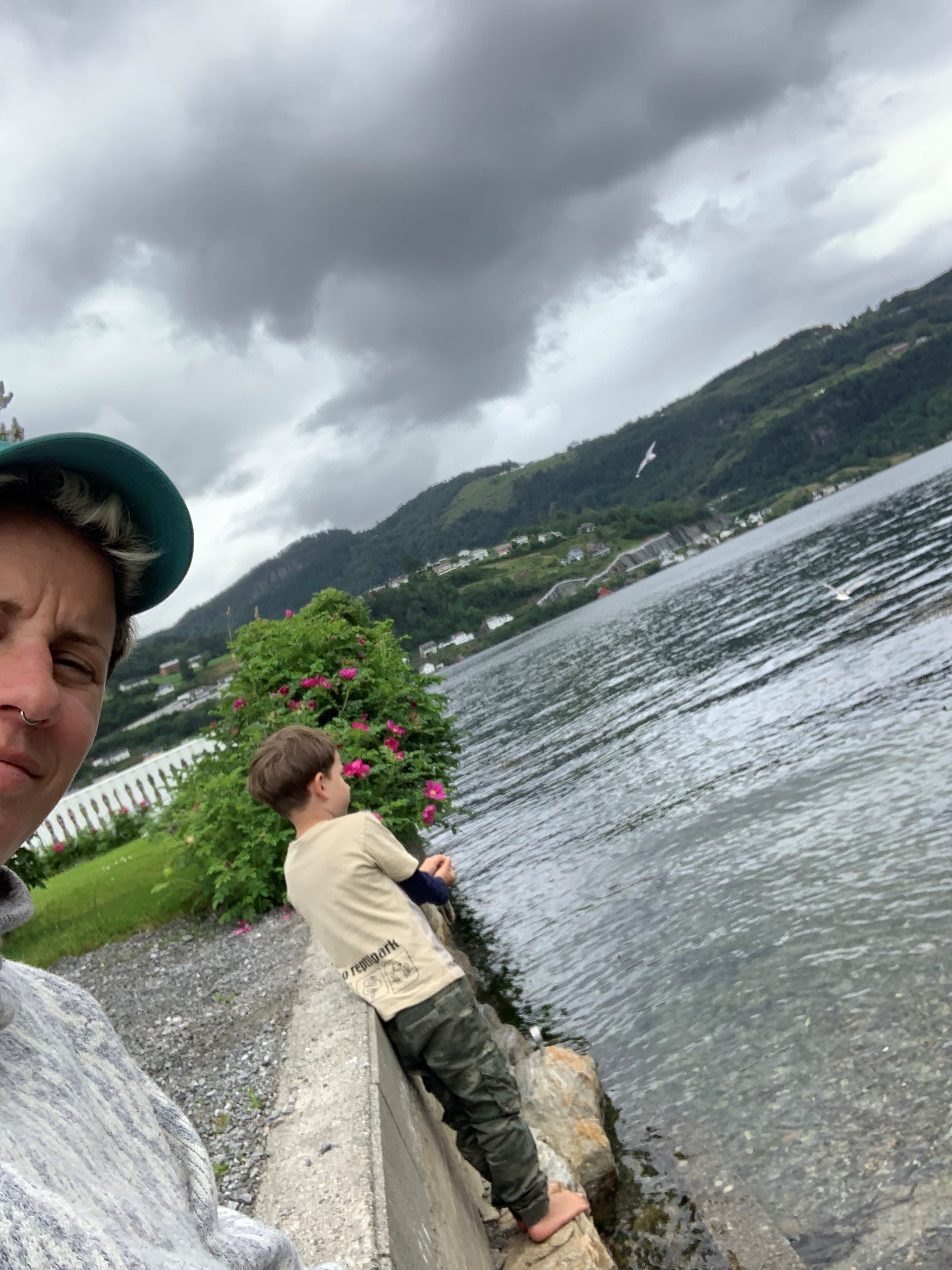 Selfie av Heidi Marie Vestrheim med caps, i bakgrunn ser vi sønnen står ved et vann med ryggen til.
