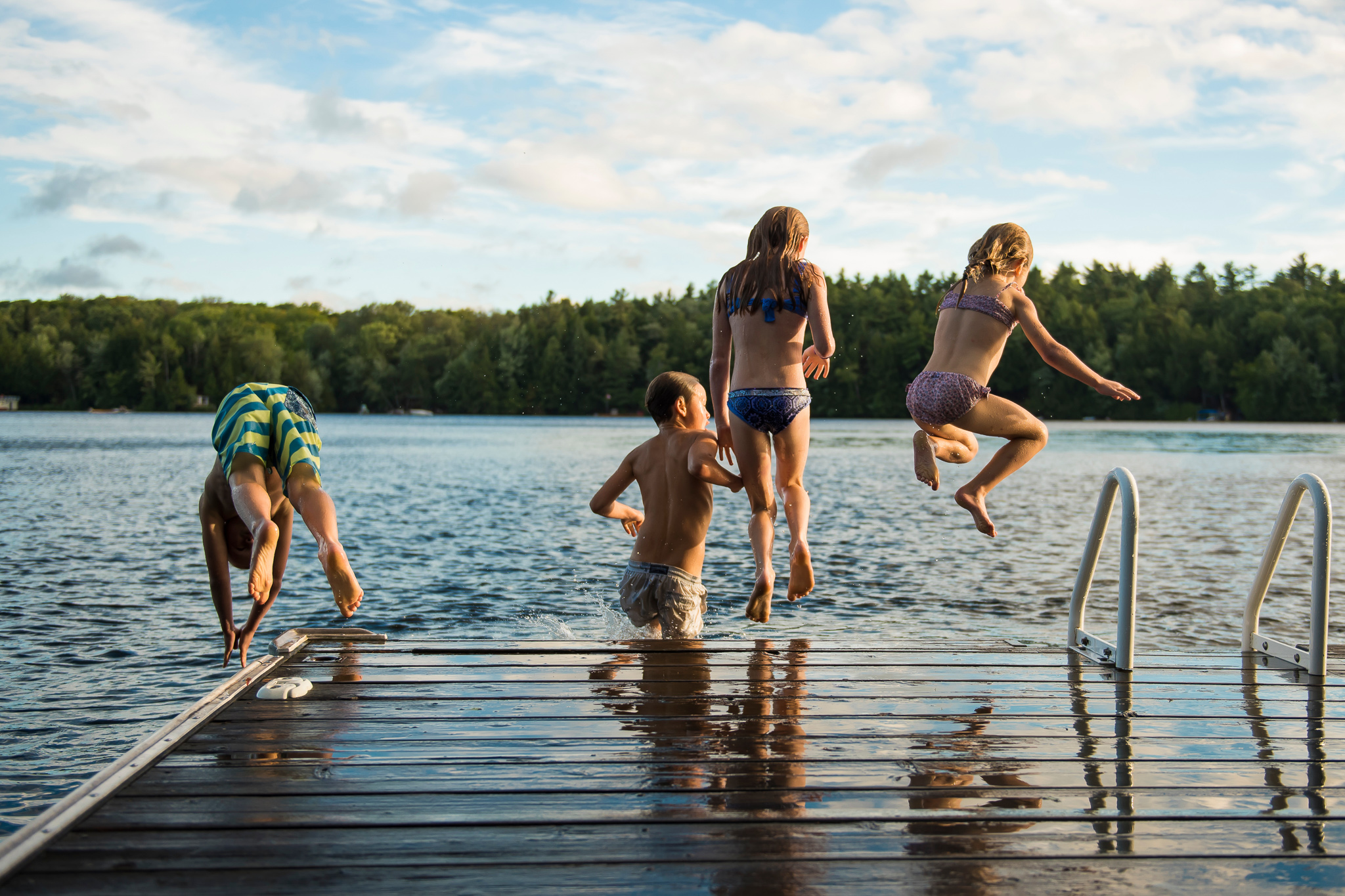 Bilde av fire barn, tre jenter og en gutt, som hopper ut i vannet fra brygge. Alle med ryggen til.