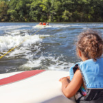 Bilde av ei jente med redningsvest som ser på barn som trekkes etter en vannleke med båten hun sitter i.