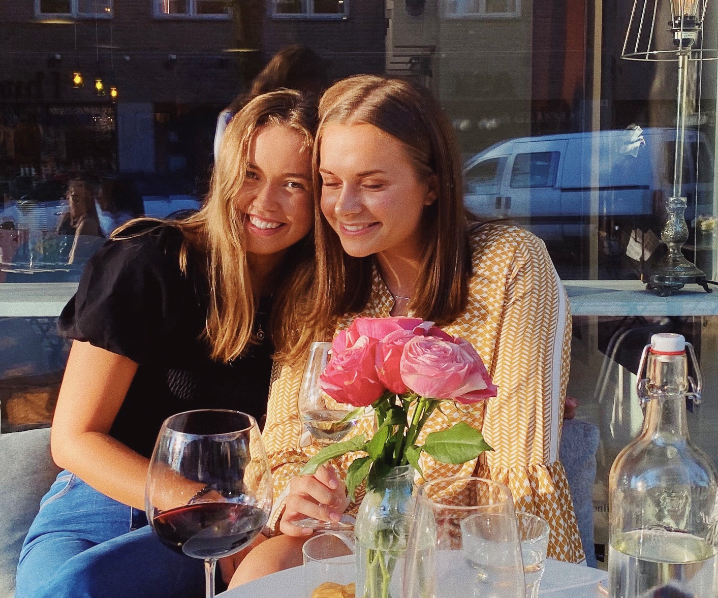 To venninner som mens de sitter lener seg mot hverandre og smiler. Foran dem er det et bord med glass og en vase med rosa blomster.
