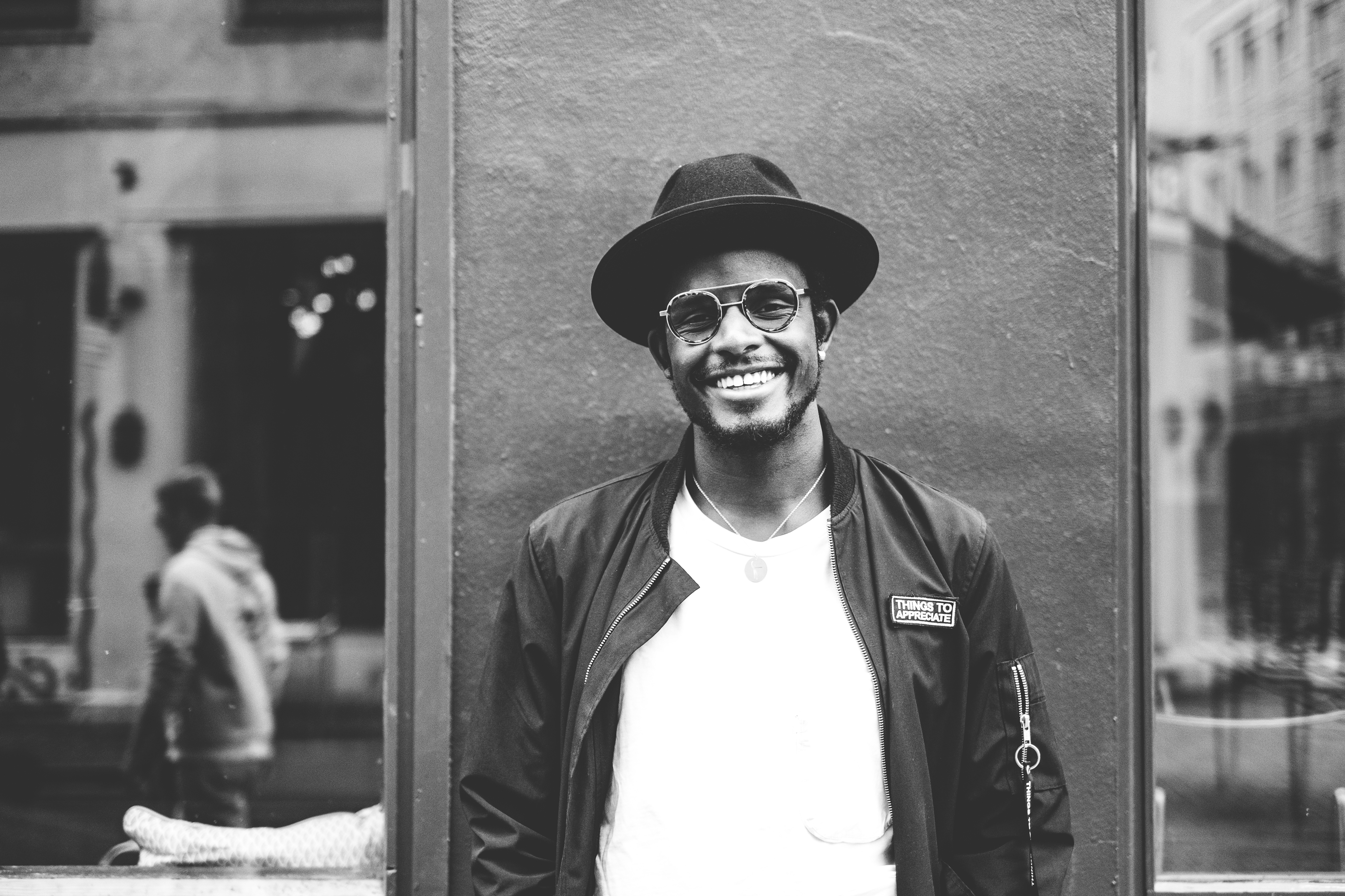Svart-hvitt bilde av en ung mann med hatt og briller, som står inntil en vegg. Han smiler bredt og ser inn i kamera.