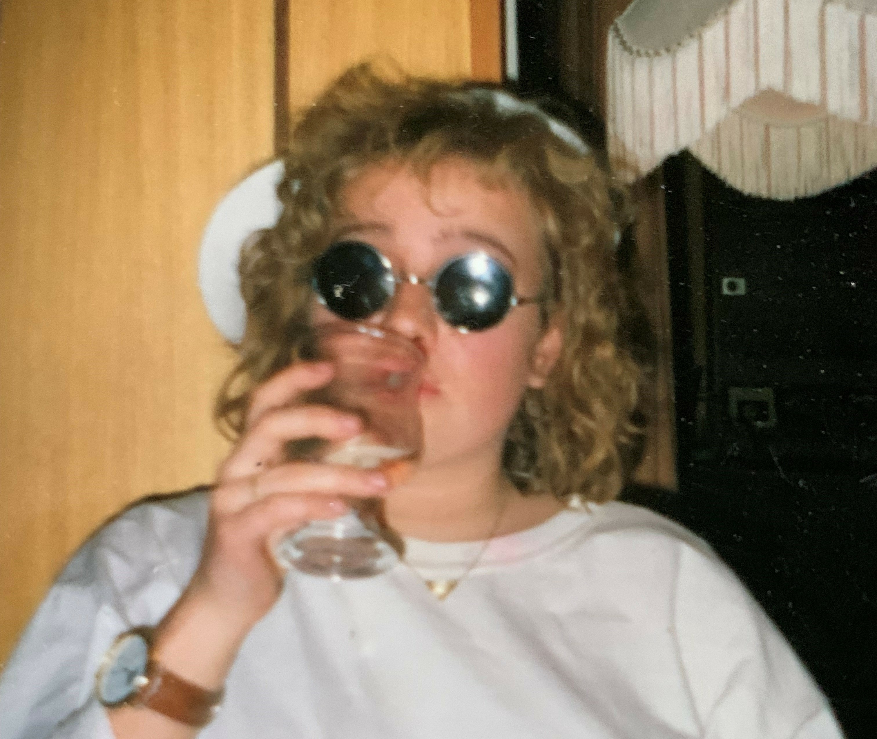 Nina Anett Lea som yngre. En ung dame lyst krøllete hår og runde solbriller. Hun tar en slurk av et glass og har blikket rettet mot kameraet.