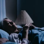 En eldre dame ligger i sengen, hun har hendene foldet over brystet og kikker opp mot taket.