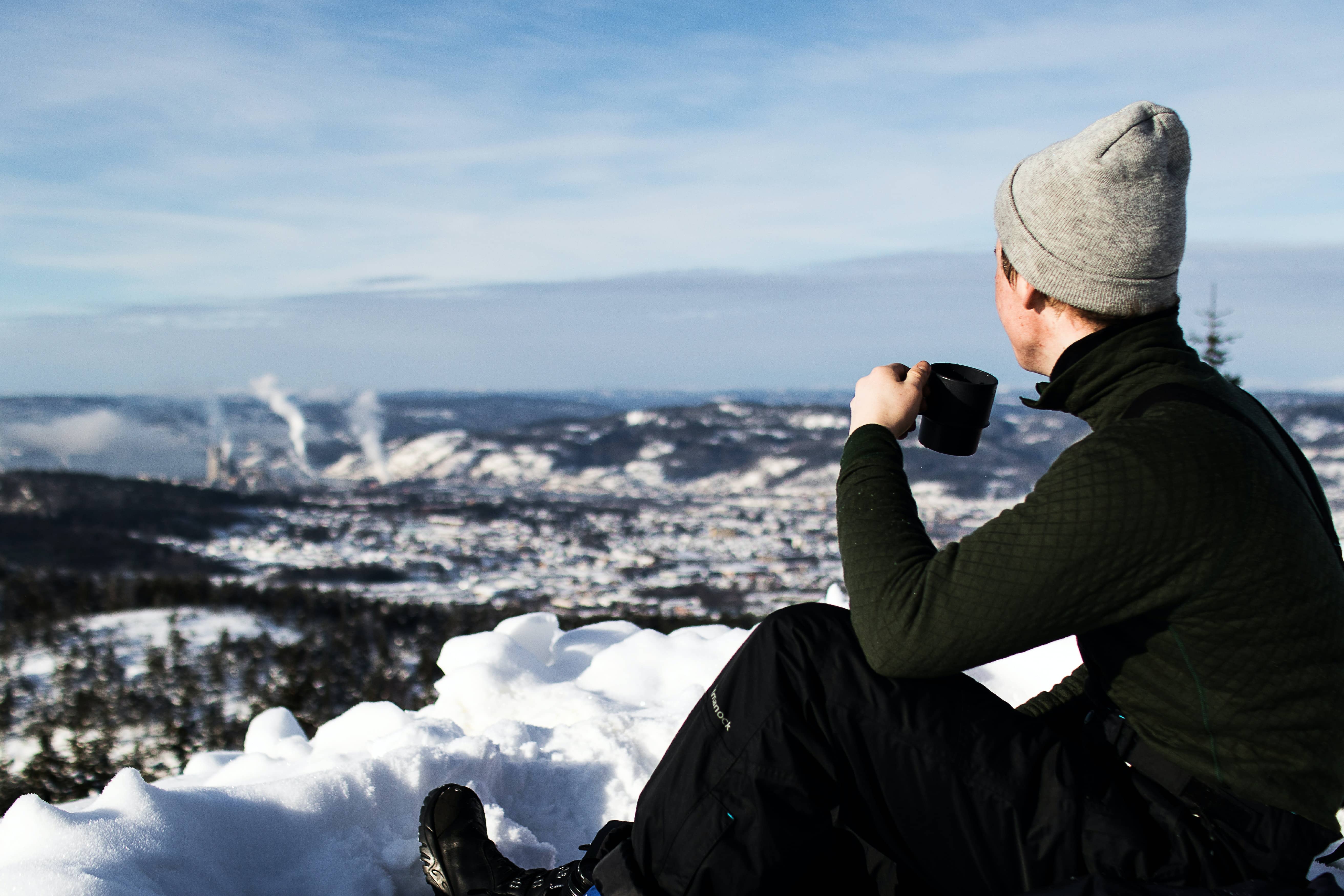 Ung mann på tur i fjellet, han sitter med en kopp kaffe og ser utover snødekket fjell.