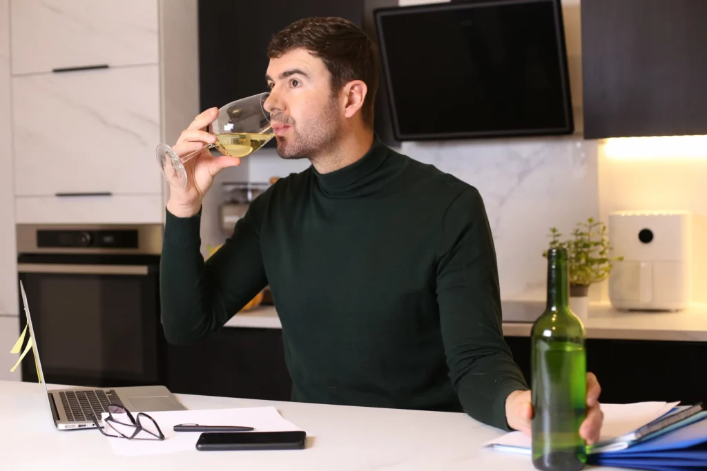 Mann sitter foran laptop på kjøkkenet. Han er på hjemmekontor og drikker alkohol fra et vinglass.