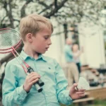 Gutt står i hage med badminton. voksne i ufokus står i bakgrunnen.
