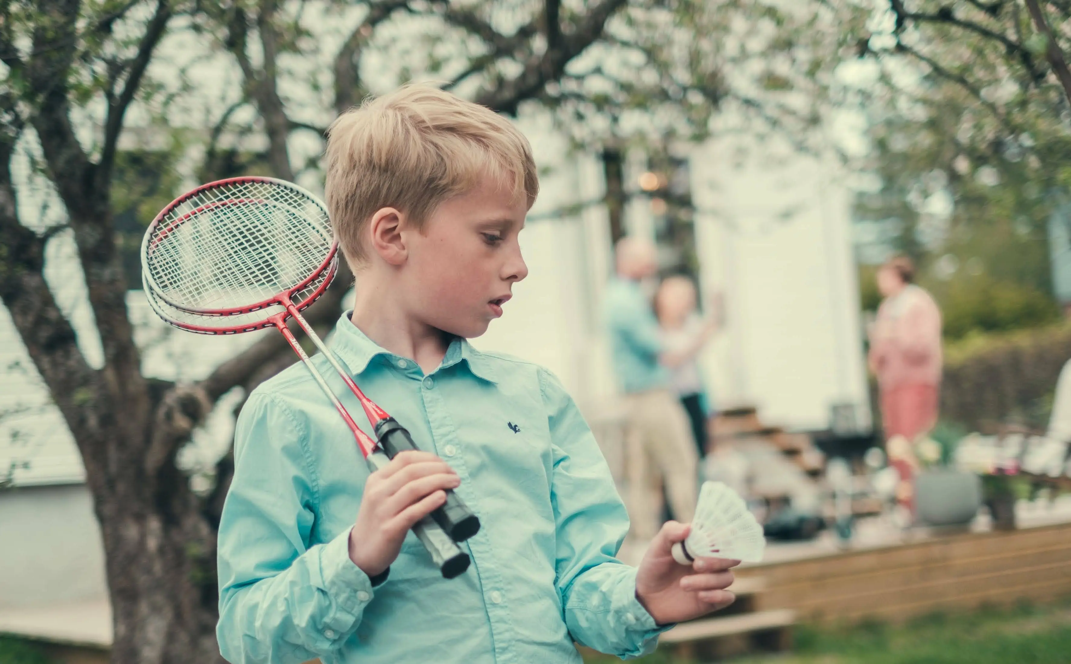 Gutt står i hage med badminton. voksne i ufokus står i bakgrunnen.